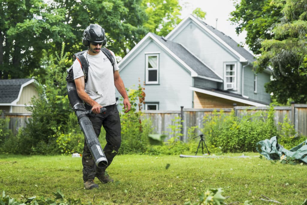Man using a leaf blower for fall clean-up in a residential yard, surrounded by greenery and a house, emphasizing professional yard maintenance by ZZ Chop Tree Care in Ottawa.