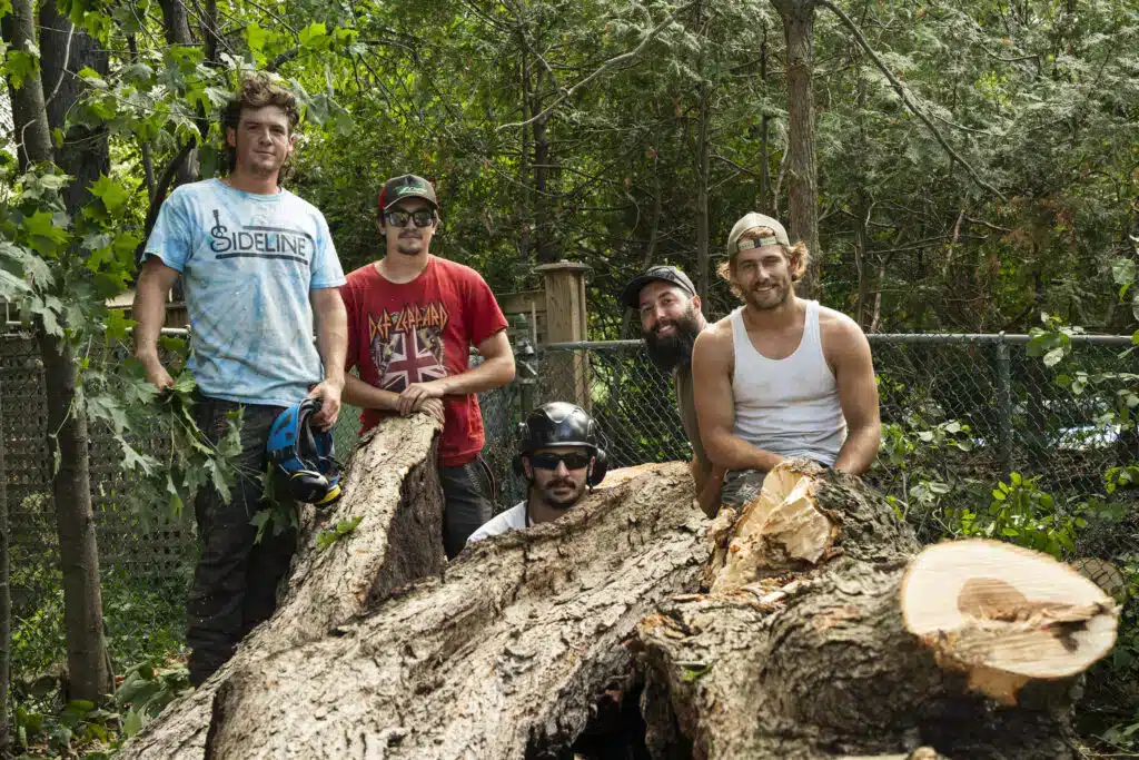 Five tree care professionals from ZZ Chop Tree Service posing with a large tree log in a wooded area, showcasing teamwork and expertise in tree removal and care.